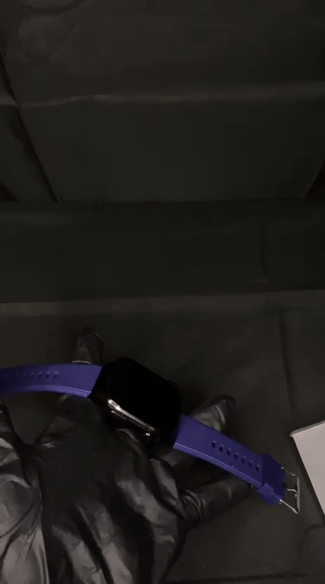 Top-down unboxing view of a black square smartwatch with a vibrant indigo purple silicone strap inside a white minimalist retail box