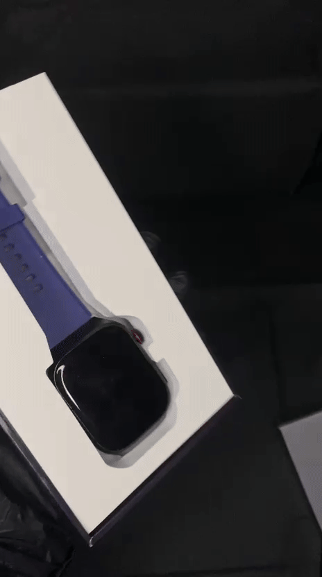 Top-down view of a black square-faced smartwatch with a deep navy blue silicone strap, resting inside its white rectangular retail packaging box.