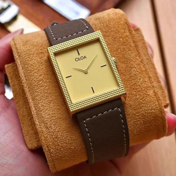 CILOA rectangular gold watch with a gold dial and smooth brown leather strap resting securely on a soft brown suede display pillow.