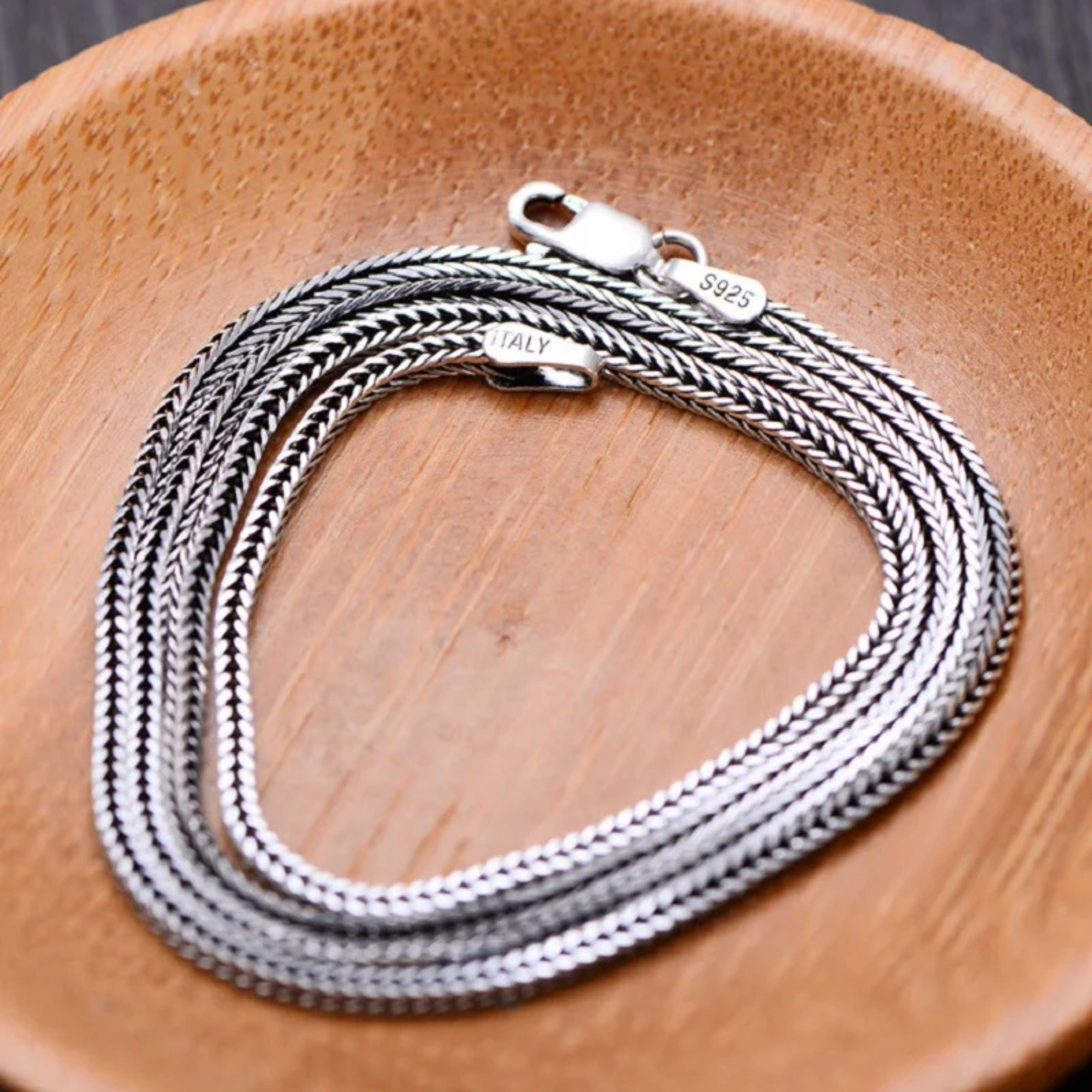 Silver woven chain necklace neatly coiled in a circle inside a warm wooden display plate.