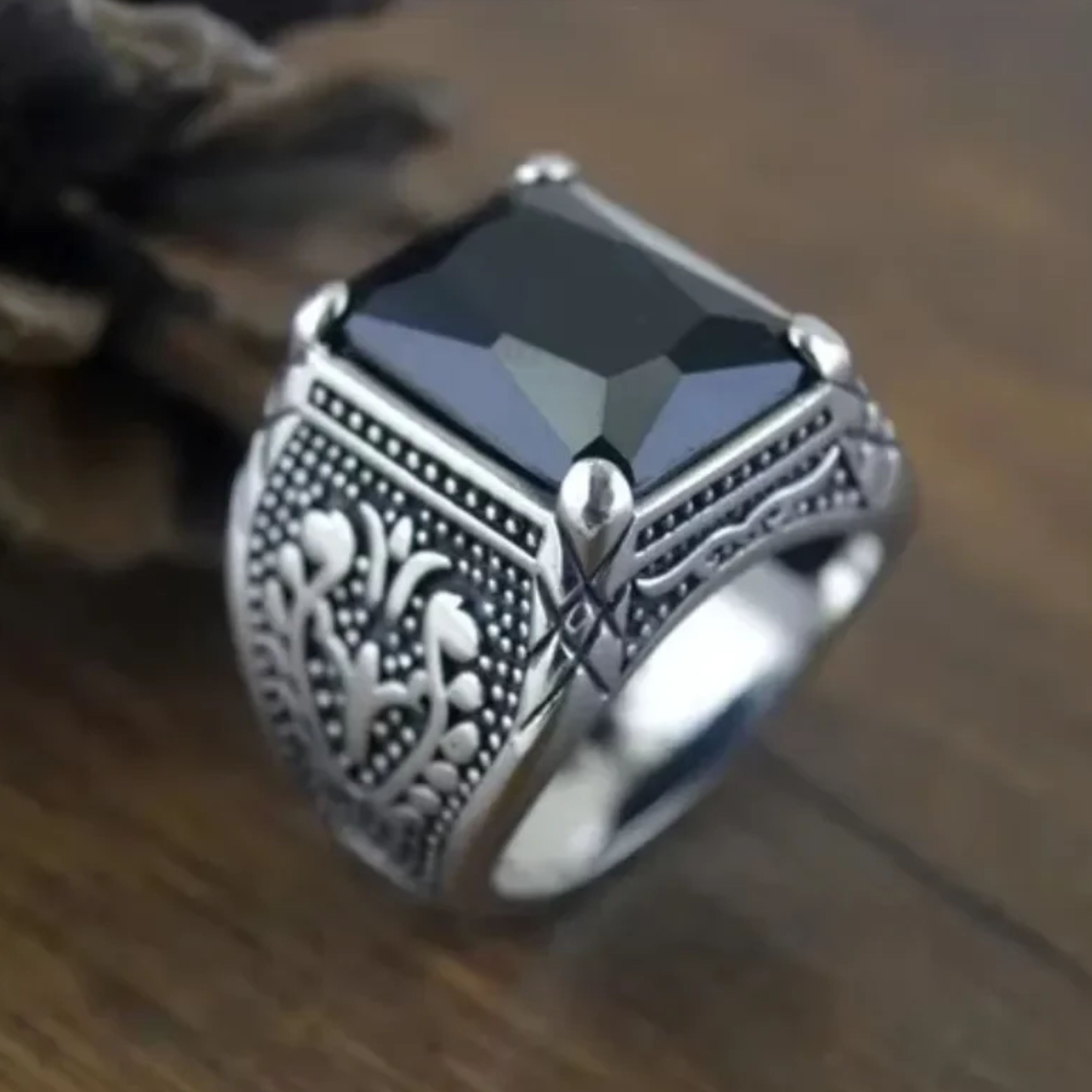 Close up of a men's silver ring with a black gemstone highlighting the detailed side engravings on dark wood.