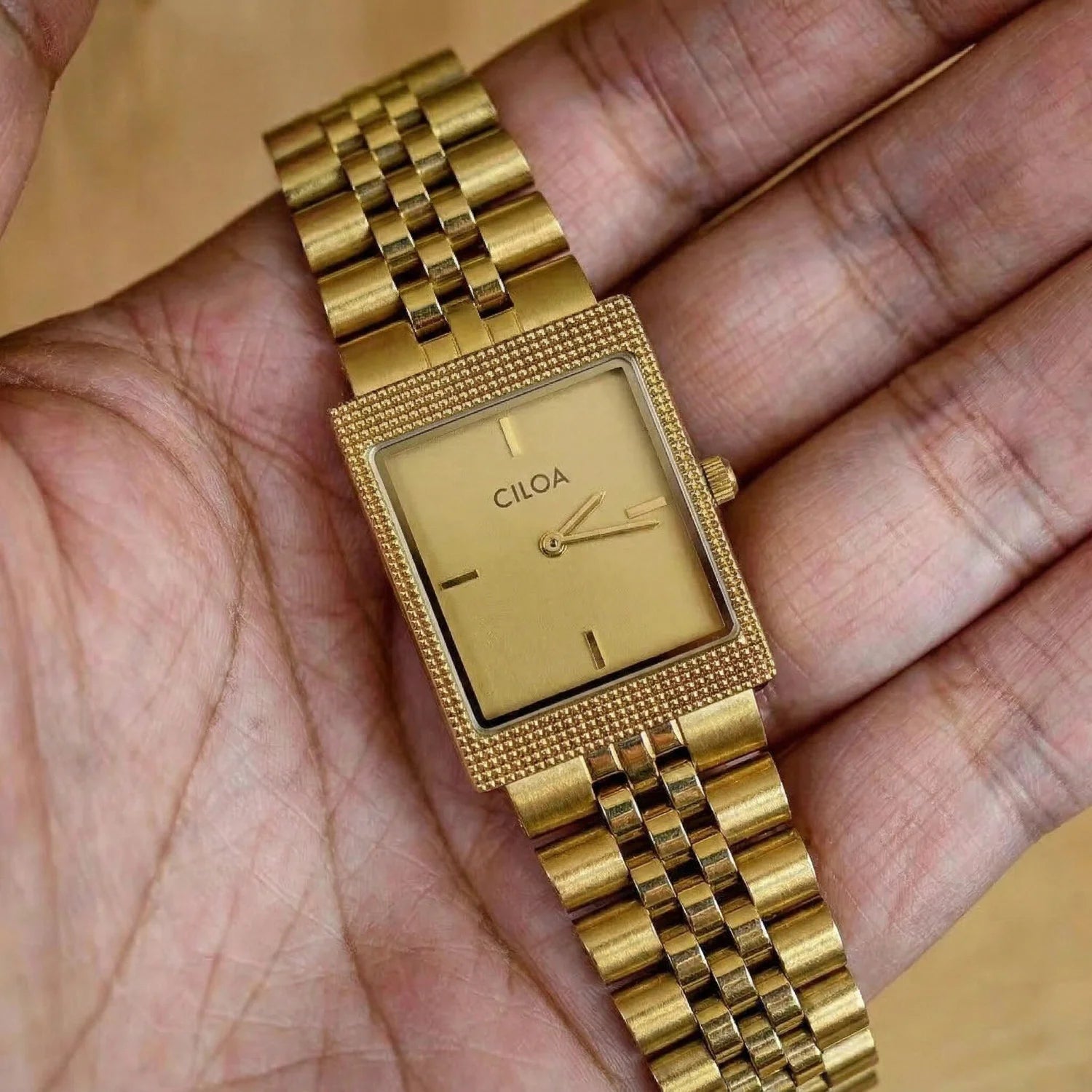 CILOA rectangular gold watch with a gold dial and metal jubilee bracelet held in the palm of a hand to showcase its elegant profile.
