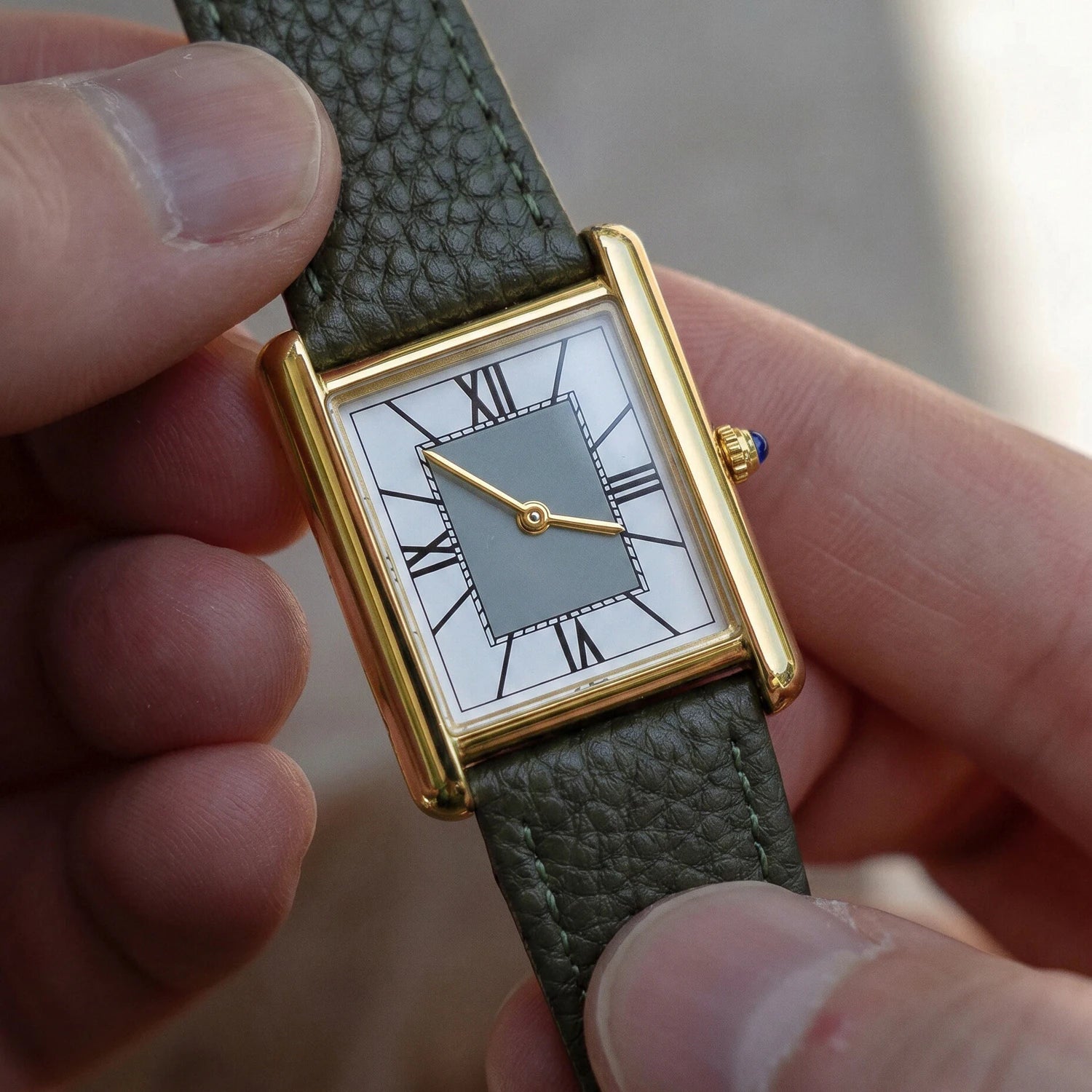 Close-up of a person holding a rectangular gold watch, highlighting the grey dial, blue crown detail, and the pebbled texture of the green leather strap.
