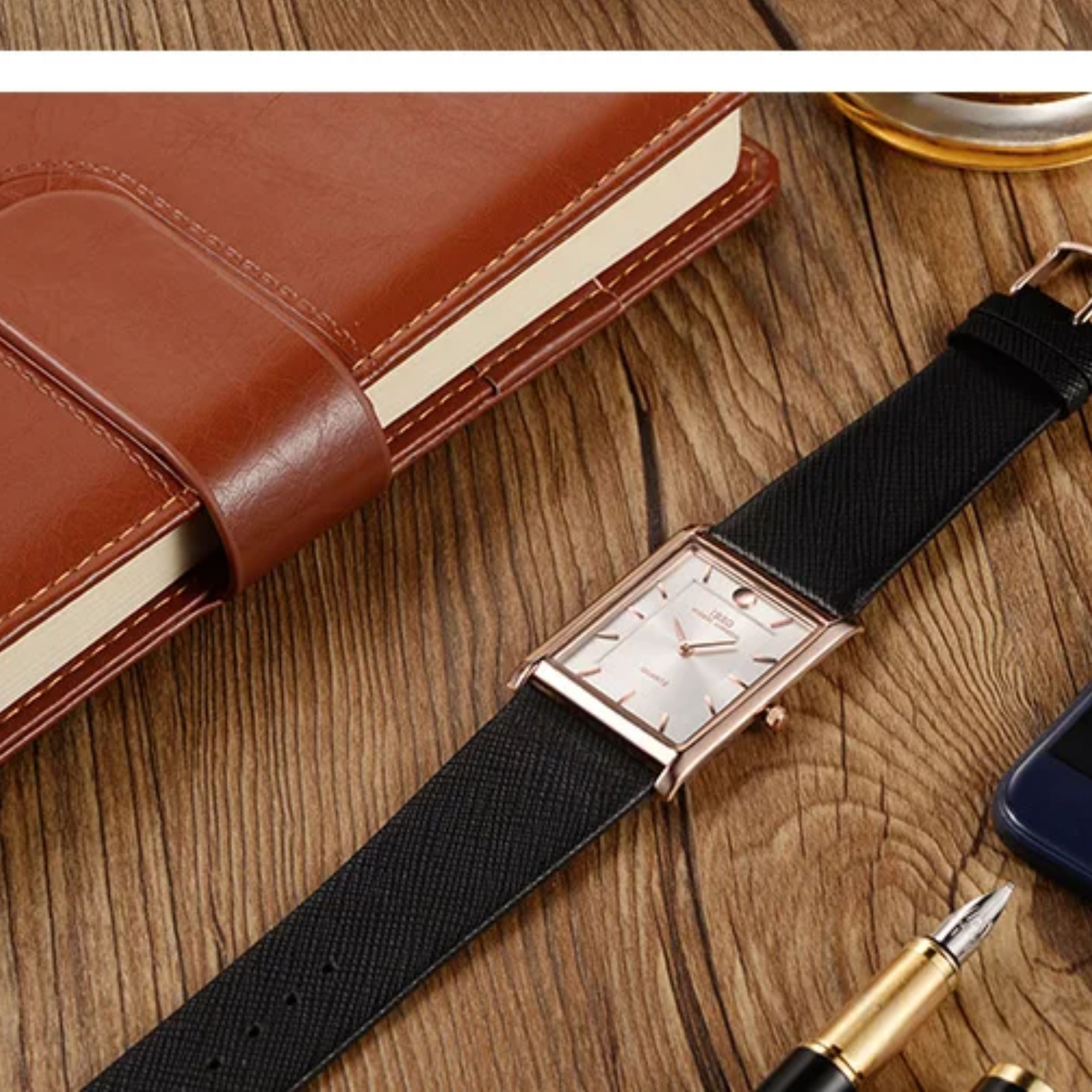 An IBSO rose gold rectangular watch with a white dial and black strap laid flat on a wooden desk next to a brown leather journal