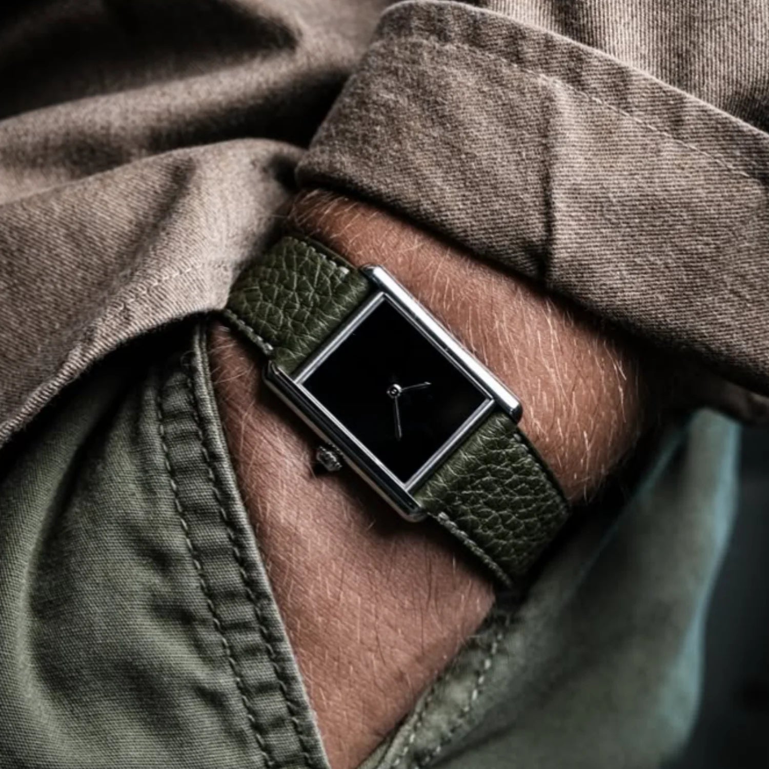 Close-up shot of a man wearing the silver CILOA tank watch with a pitch black minimalist dial and an olive green textured leather strap