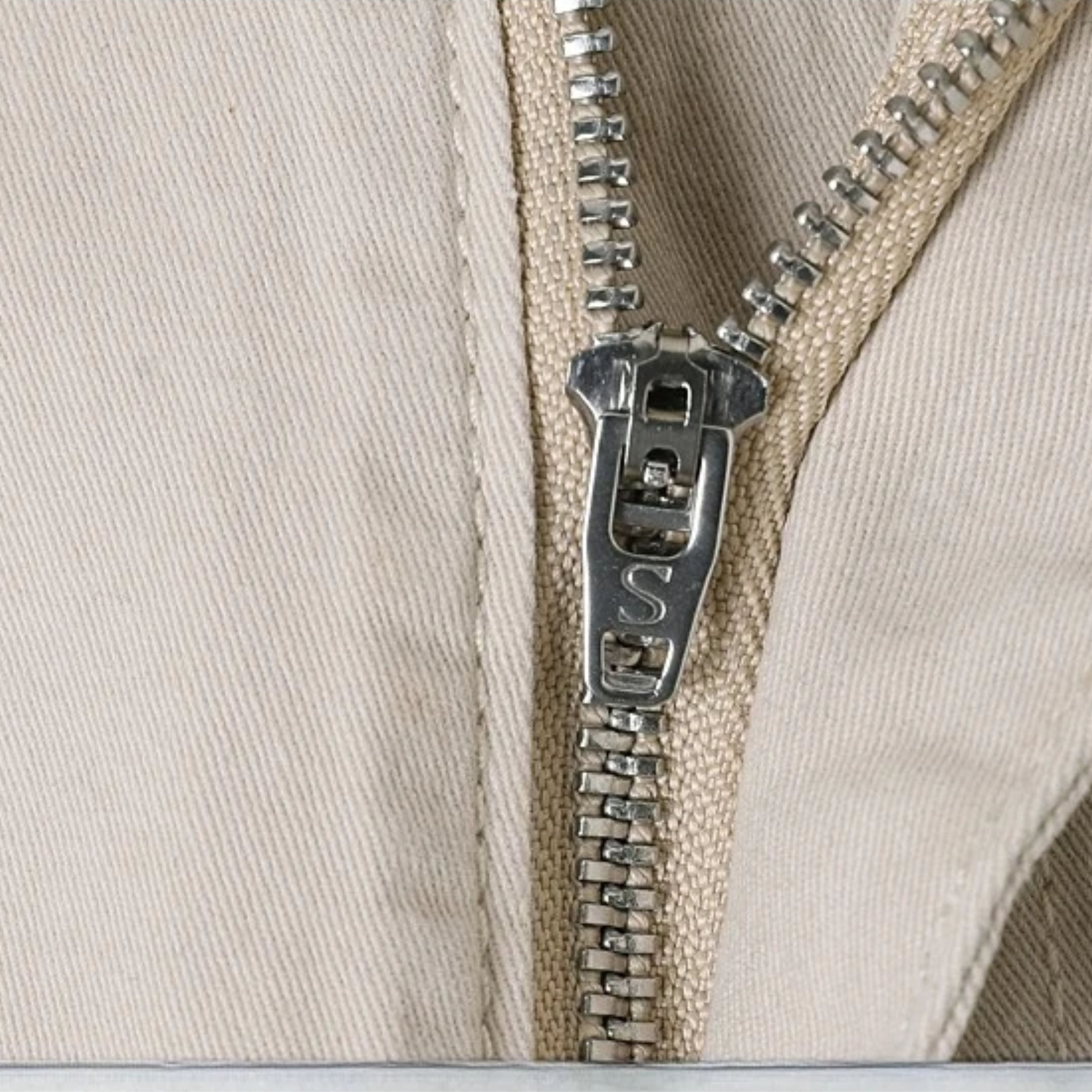 A technical macro close-up highlighting the secure metallic single-glide zipper fly closure and rugged interlock tracks on the light-wash casual trousers