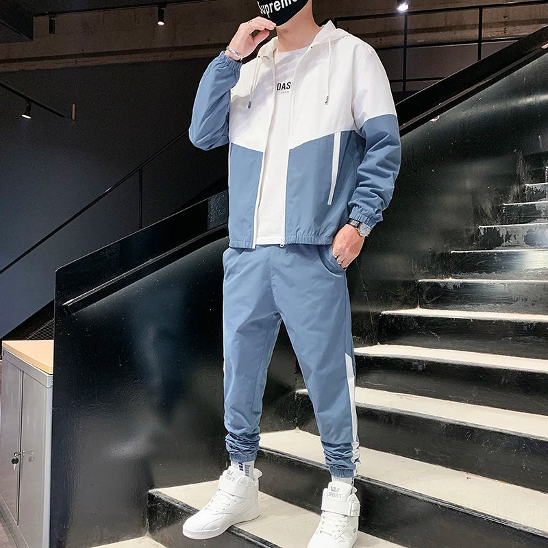A male model posing on modern stairs wearing the light blue and white colorblock mandarin collar hooded jacket and matching joggers set