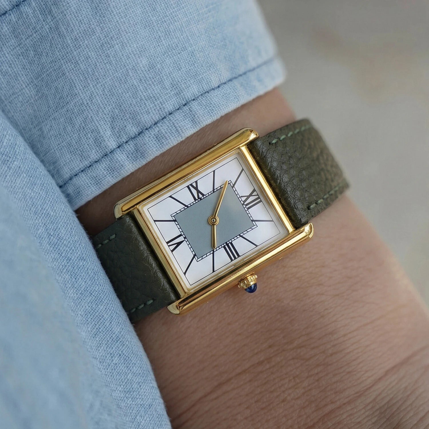 Model wearing a vintage style rectangular gold watch with an olive green leather strap, paired with a light blue dress shirt.