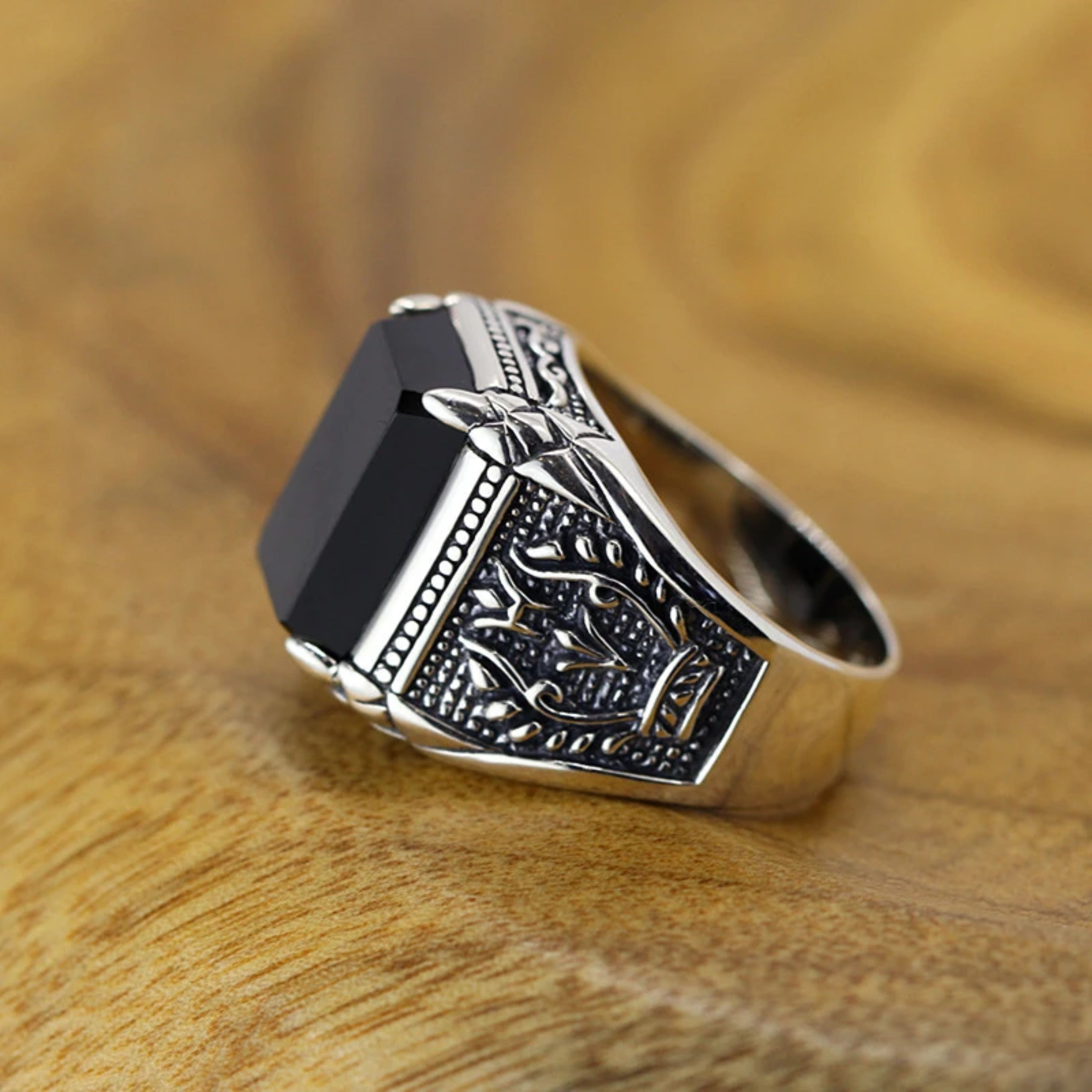 Silver signet ring with engraved floral sides and a black stone resting on a warm wooden surface.
