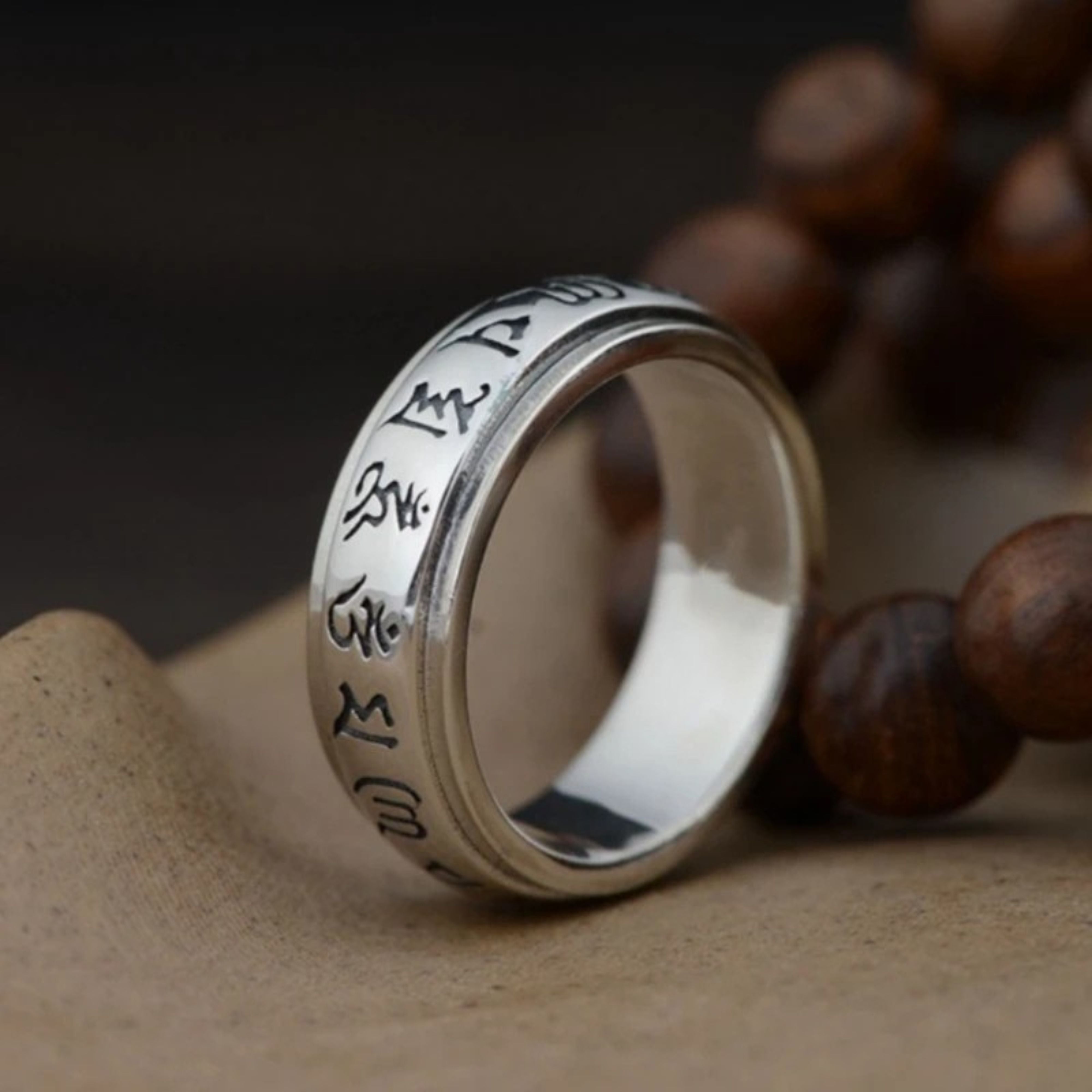 Silver Buddhist prayer ring standing upright on a textured surface with blurred prayer beads in the dark background.