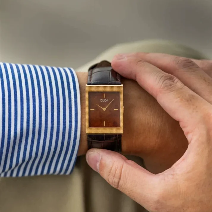 Lifestyle shot of a man's wrist wearing a rectangular gold tank-style watch with a brown dial, resting his hand over his knee.