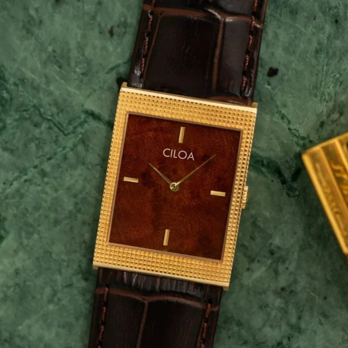CILOA rectangular gold watch with a textured brown dial and crocodile leather strap resting flat on a dark green textured surface.