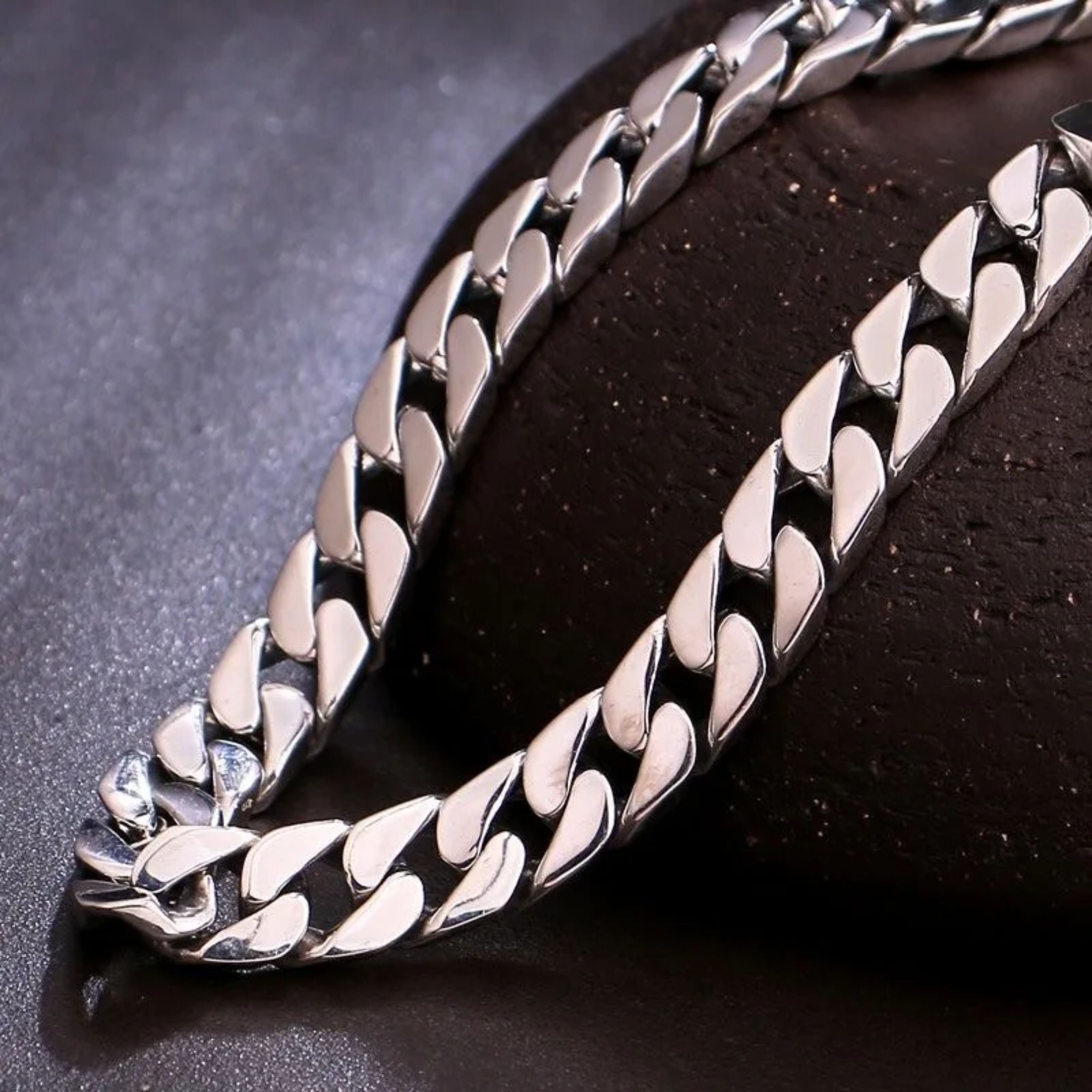 Macro shot highlighting the flat, tightly interlocked links of a heavy silver curb chain on a dark surface.