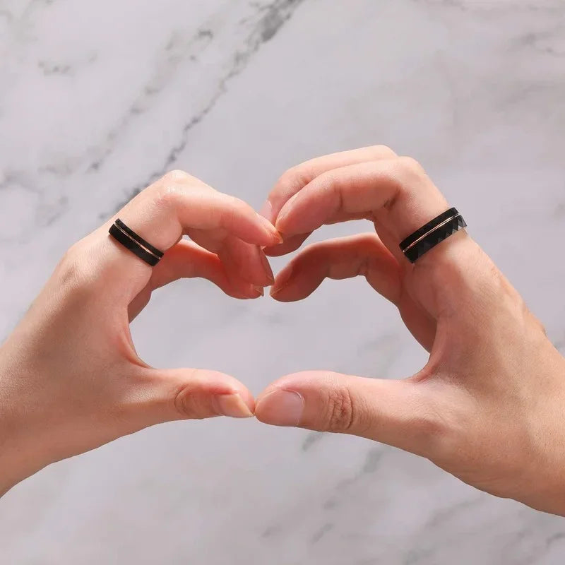 Two hands making a heart shape, displaying matching Tigrade black faceted tungsten promise rings with rose gold accents on a marble background