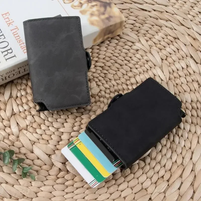 Two black minimalist pop-up card wallets arranged as a lifestyle flat lay on a textured woven straw mat next to a book