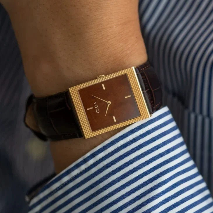 Close-up wrist shot of a men's rectangular gold watch with a brown dial worn elegantly under a dark striped shirt cuff.