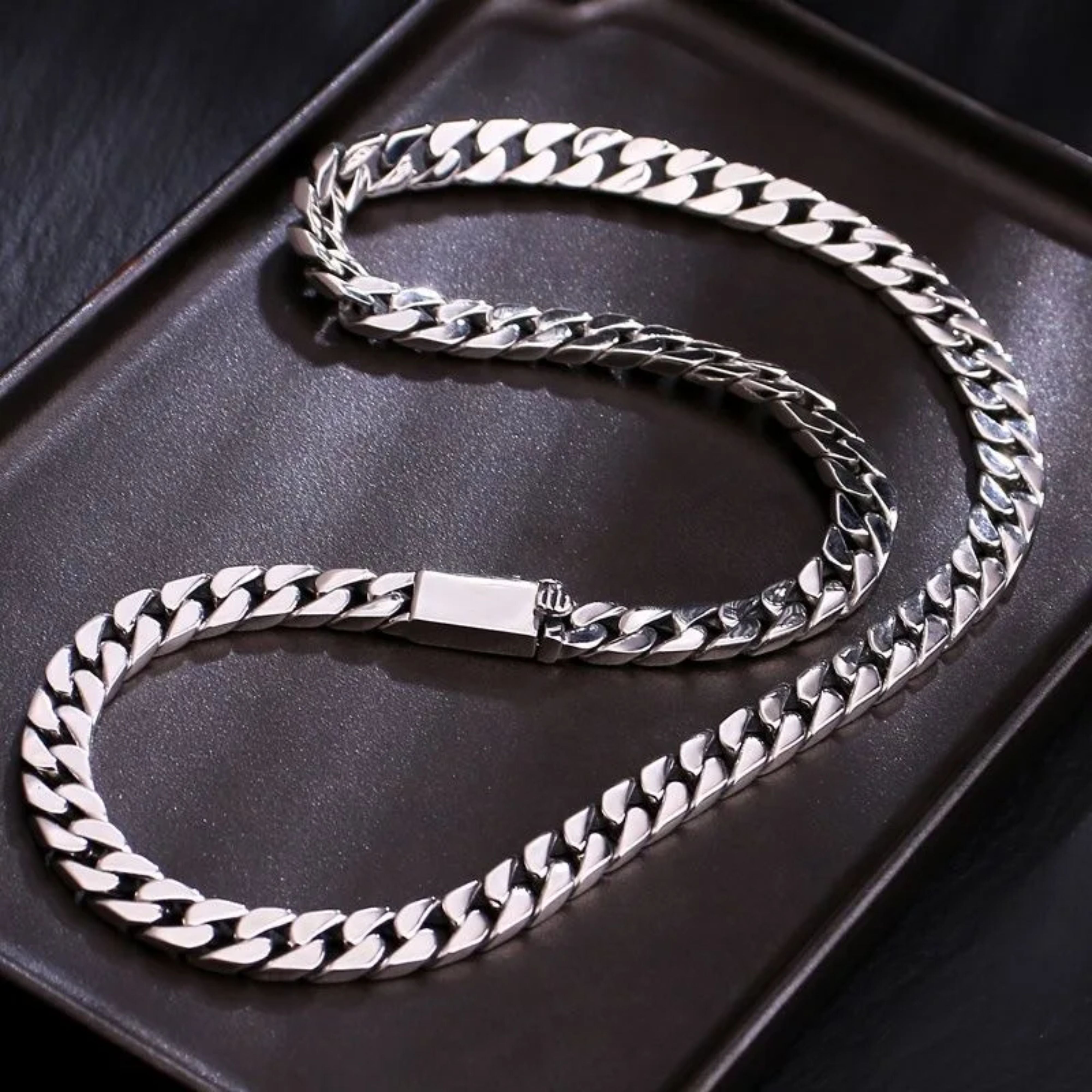 Men's silver Cuban chain necklace arranged in a curved S-shape on a dark brown display tray.