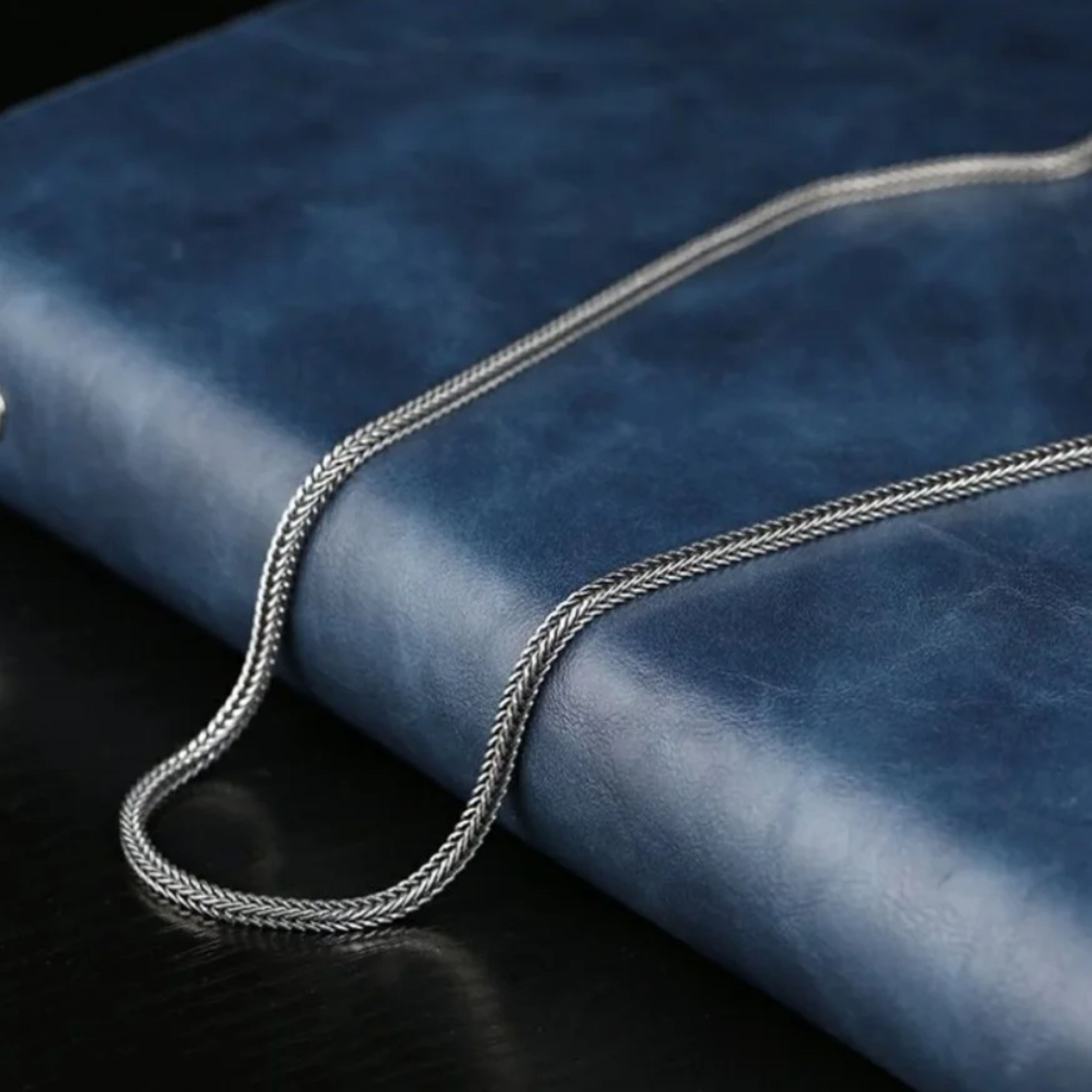 Men's silver wheat chain necklace draped elegantly across a blue leather-bound surface.