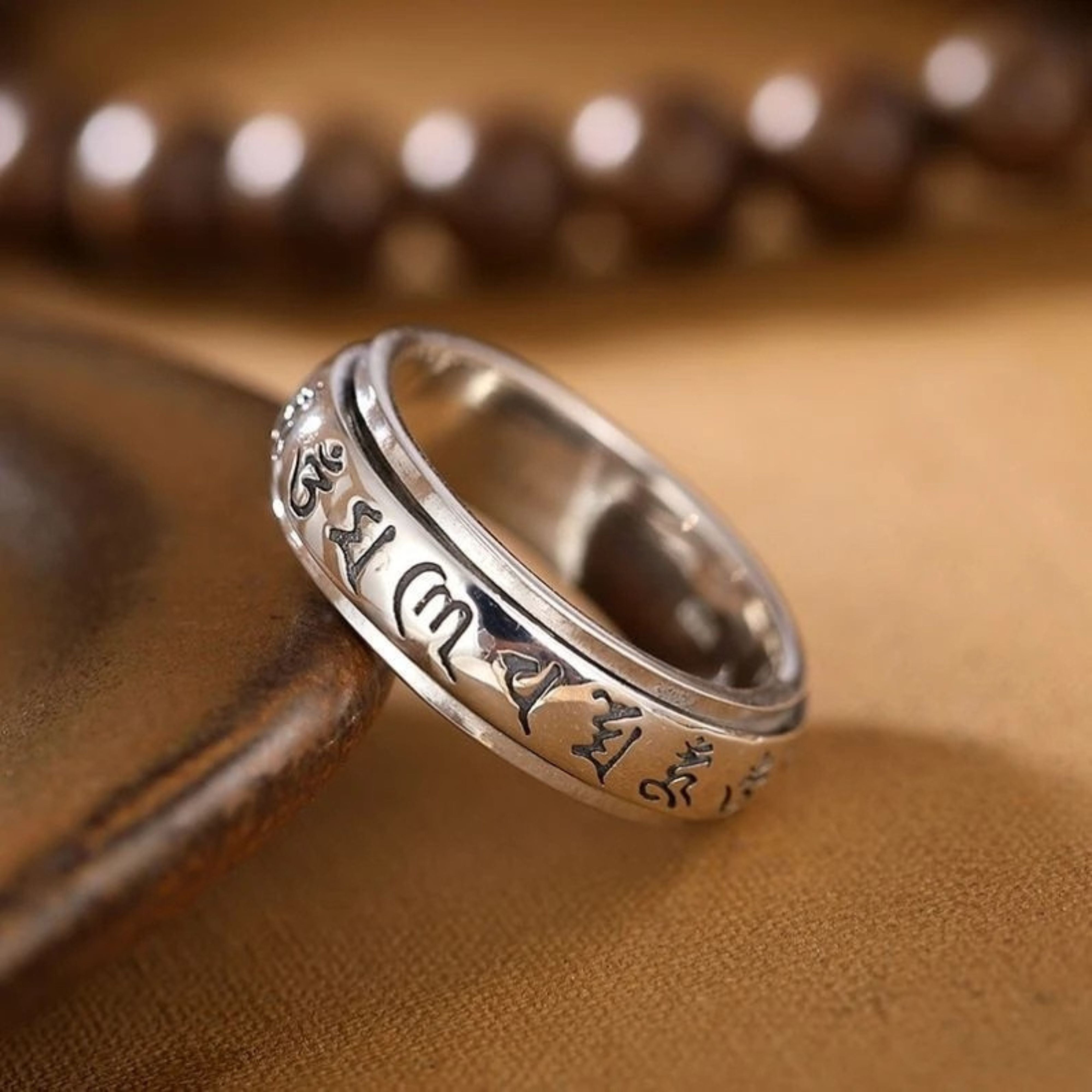 Silver Tibetan mantra meditation ring resting on a warm leather surface next to traditional wooden prayer beads.