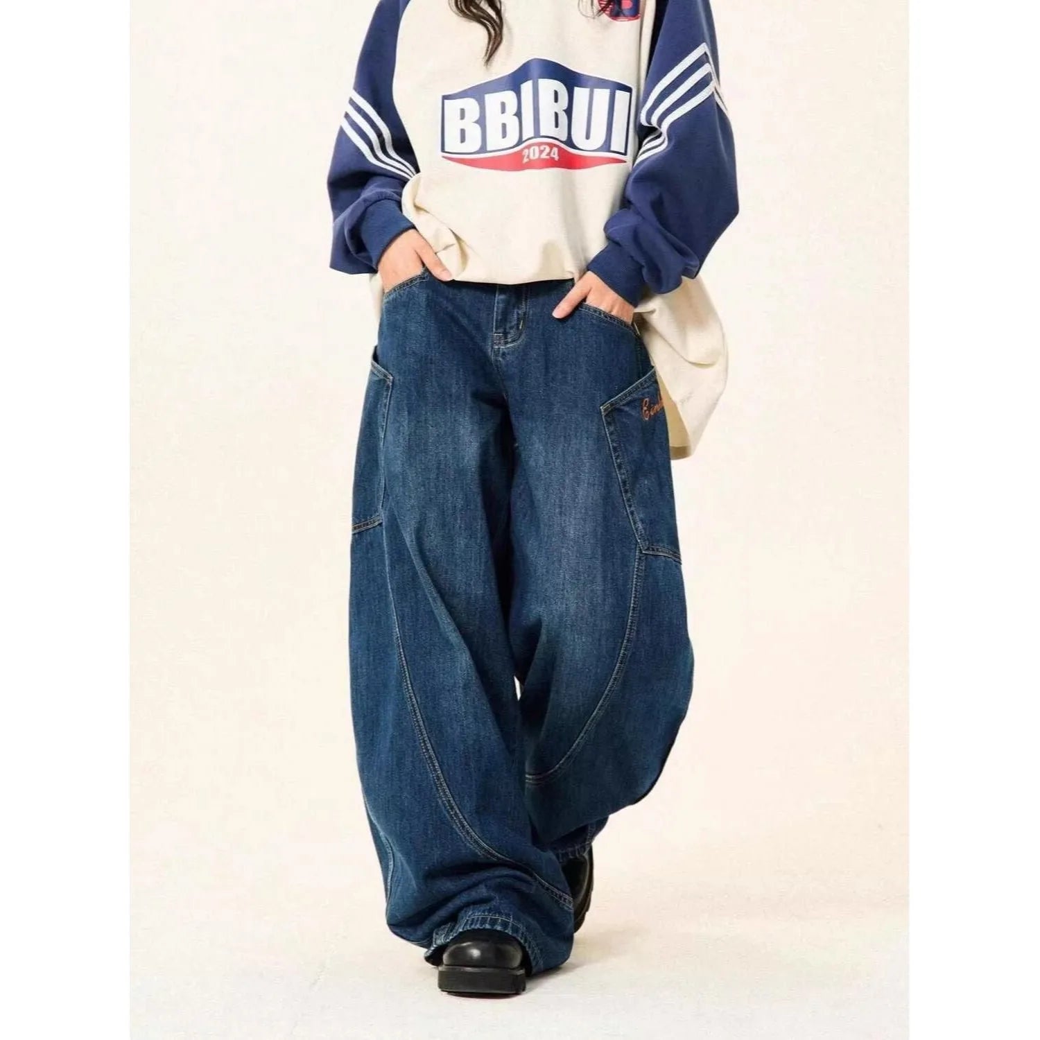 Model styling a casual streetwear outfit with a navy varsity sweatshirt and blue baggy jeans featuring curved stitching panel lines