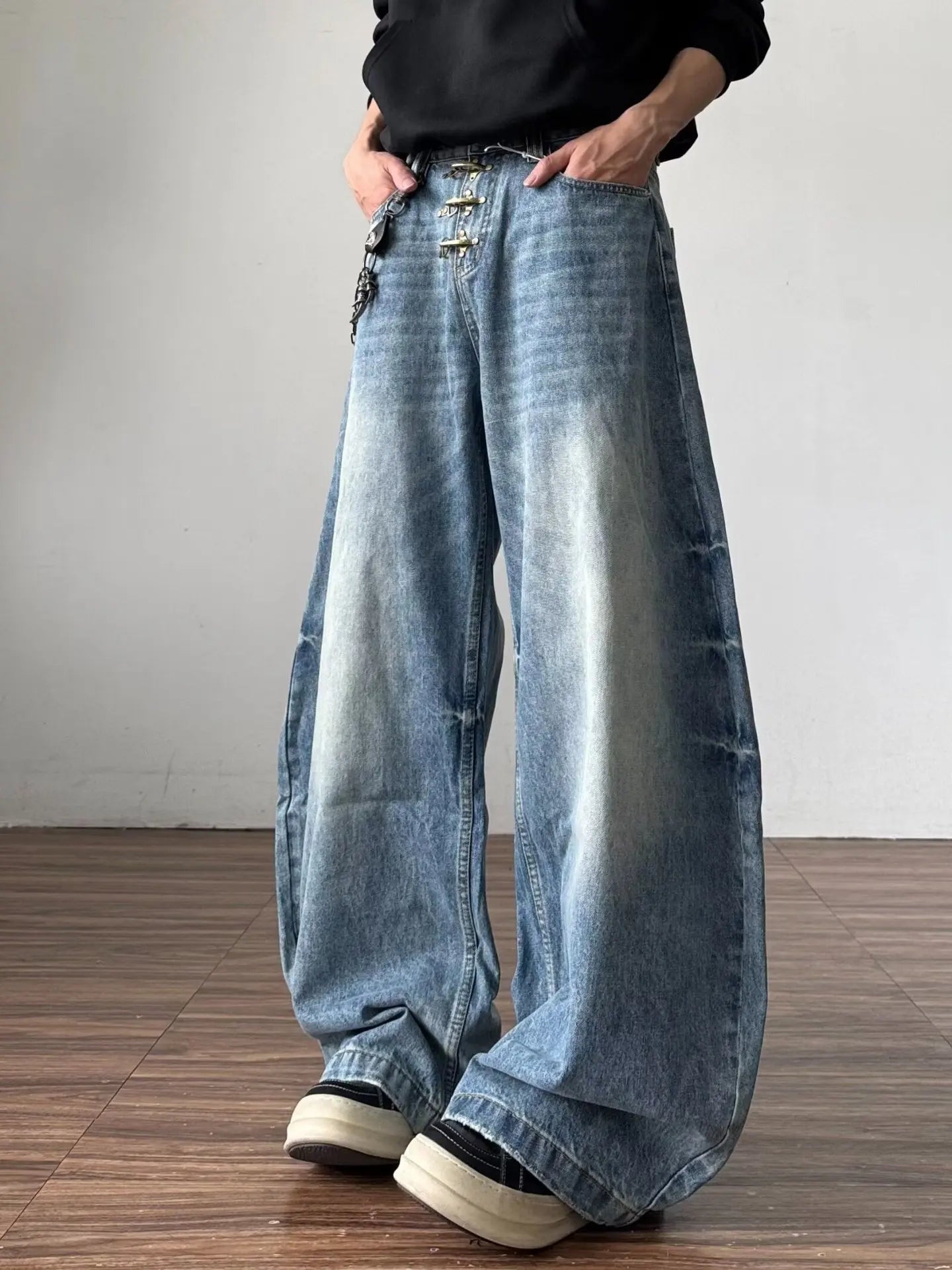 Three-quarter view of a model styling light wash asymmetrical multi-button baggy jeans by THE CREW on wooden flooring