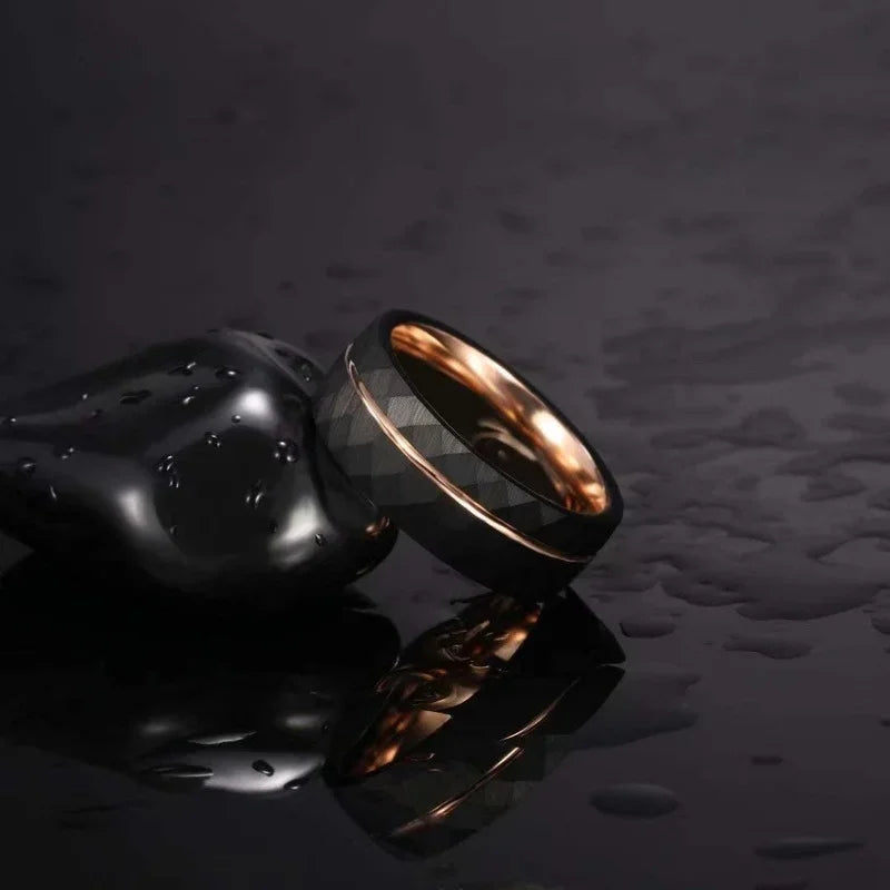 Two Tigrade hammered black tungsten rings with rose gold interiors stacked on a dark, wet reflective surface next to smooth black stones